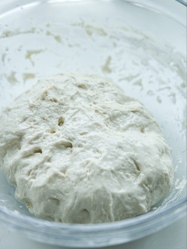 The Perfect Fluffy Focaccia Rossa Recipe - So Easy! 16 A close-up of risen bread dough with a slightly sticky texture, resting in a clear glass bowl. The dough has visible air bubbles and an uneven surface, indicating it has been proofed.
