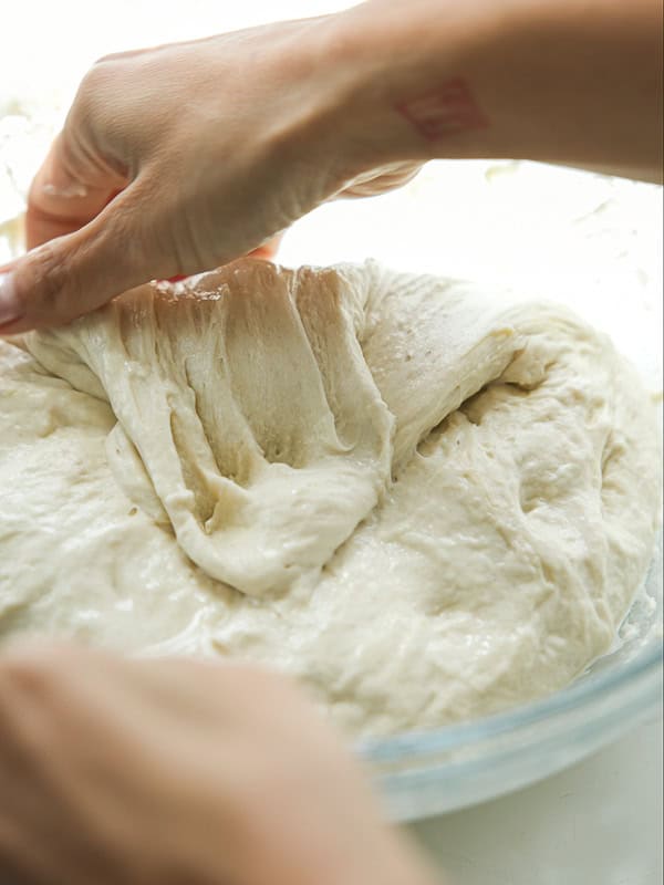 The Perfect Fluffy Focaccia Rossa Recipe - So Easy! 17 A close-up of hands stretching and folding a soft, sticky dough in a glass bowl, preparing it for baking.