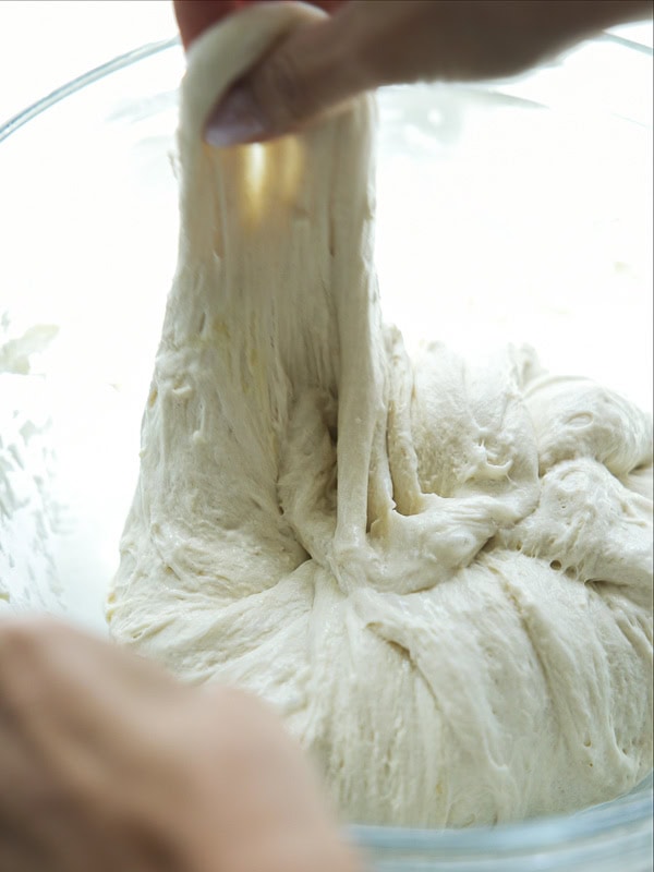 The Perfect Fluffy Focaccia Rossa Recipe - So Easy! 18 A close-up of hands stretching and pulling soft, sticky bread dough in a glass bowl.