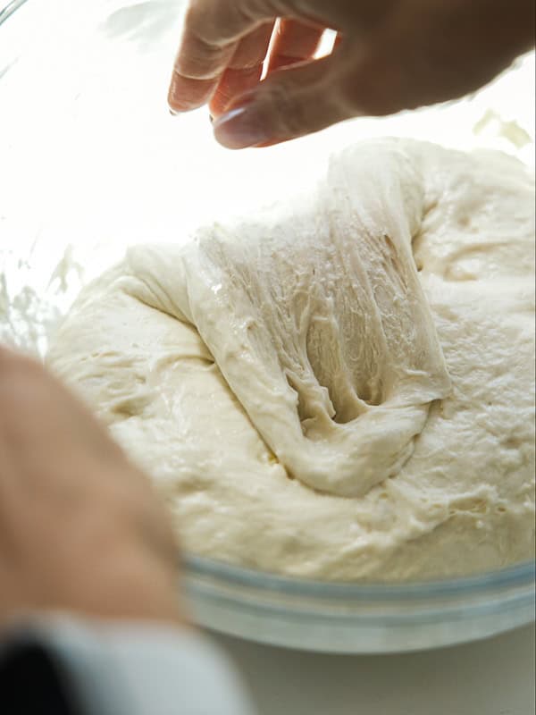 The Perfect Fluffy Focaccia Rossa Recipe - So Easy! 19 A close-up of hands stretching and folding soft, risen bread dough in a glass bowl.