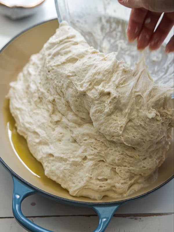 The Perfect Fluffy Focaccia Rossa Recipe - So Easy! 23 A hand transfers soft, airy bread dough from a glass bowl into a blue enameled baking dish with a layer of olive oil on the bottom.