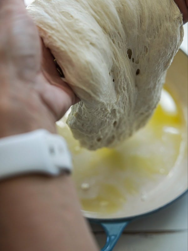 The Perfect Fluffy Focaccia Rossa Recipe - So Easy! 24 A person with a white watch stretches soft, airy bread dough over a blue dish filled with olive oil.