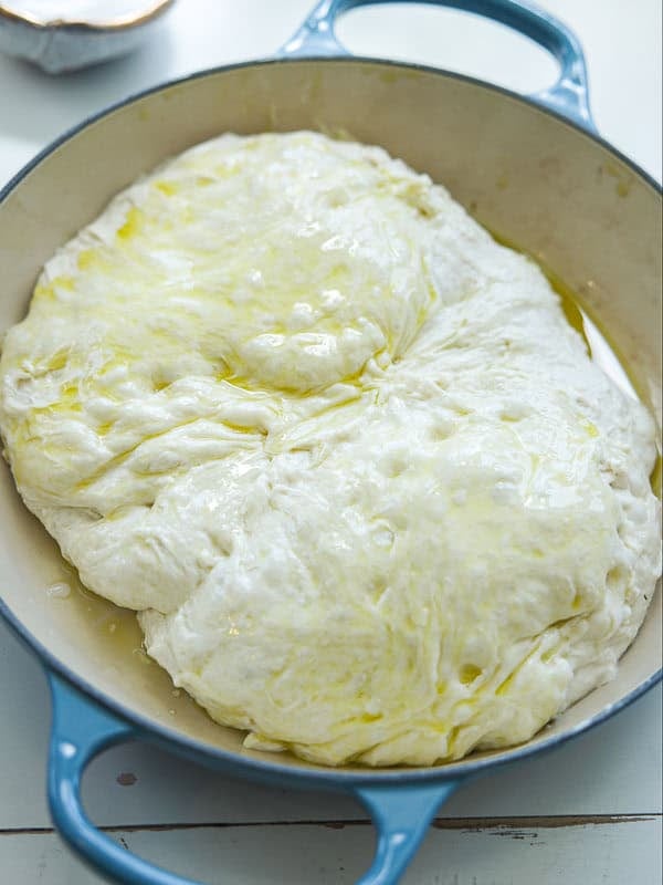 The Perfect Fluffy Focaccia Rossa Recipe - So Easy! 25 A round blue baking dish filled with risen, unbaked focaccia dough, glistening with olive oil on the surface.