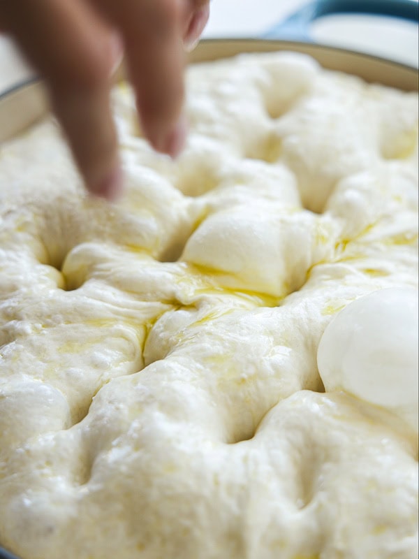 The Perfect Fluffy Focaccia Rossa Recipe - So Easy! 26 A close-up of a hand pressing fingers into soft, risen focaccia dough in a pan, with pools of olive oil visible on the surface.