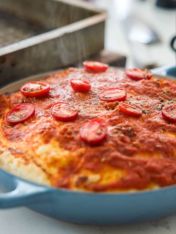 A close-up of a baked focaccia rossa in a blue pan, topped with tomato sauce, herbs, and sliced cherry tomatoes. Steam is rising, indicating the pizza is hot and freshly made.