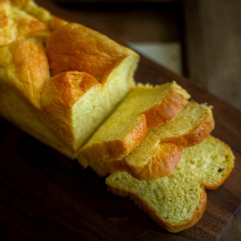 The Korean Vegan: HomeMade 44 A loaf of golden, fluffy bread with three slices cut, resting on a dark wooden surface—perfect for enjoying while you browse your next Book PreOrder. The bread appears soft and airy, with a slightly shiny, browned crust.