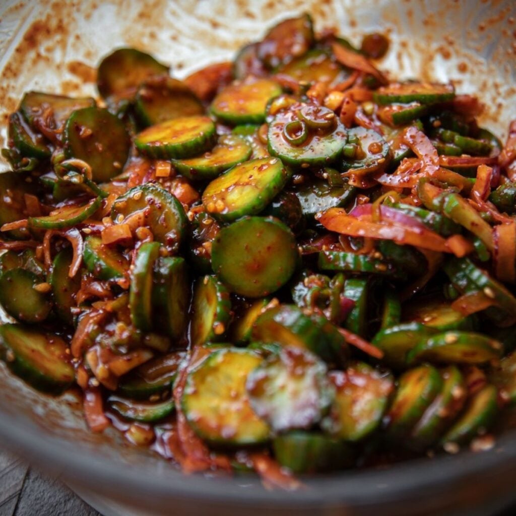 The Korean Vegan: HomeMade 50 A close-up of a bowl filled with spicy cucumber salad—sliced cucumbers, shredded carrots, red onions, and chili sauce—all coated in a flavorful dressing. Perfect for your next meal or as inspiration from our latest Book PreOrder.