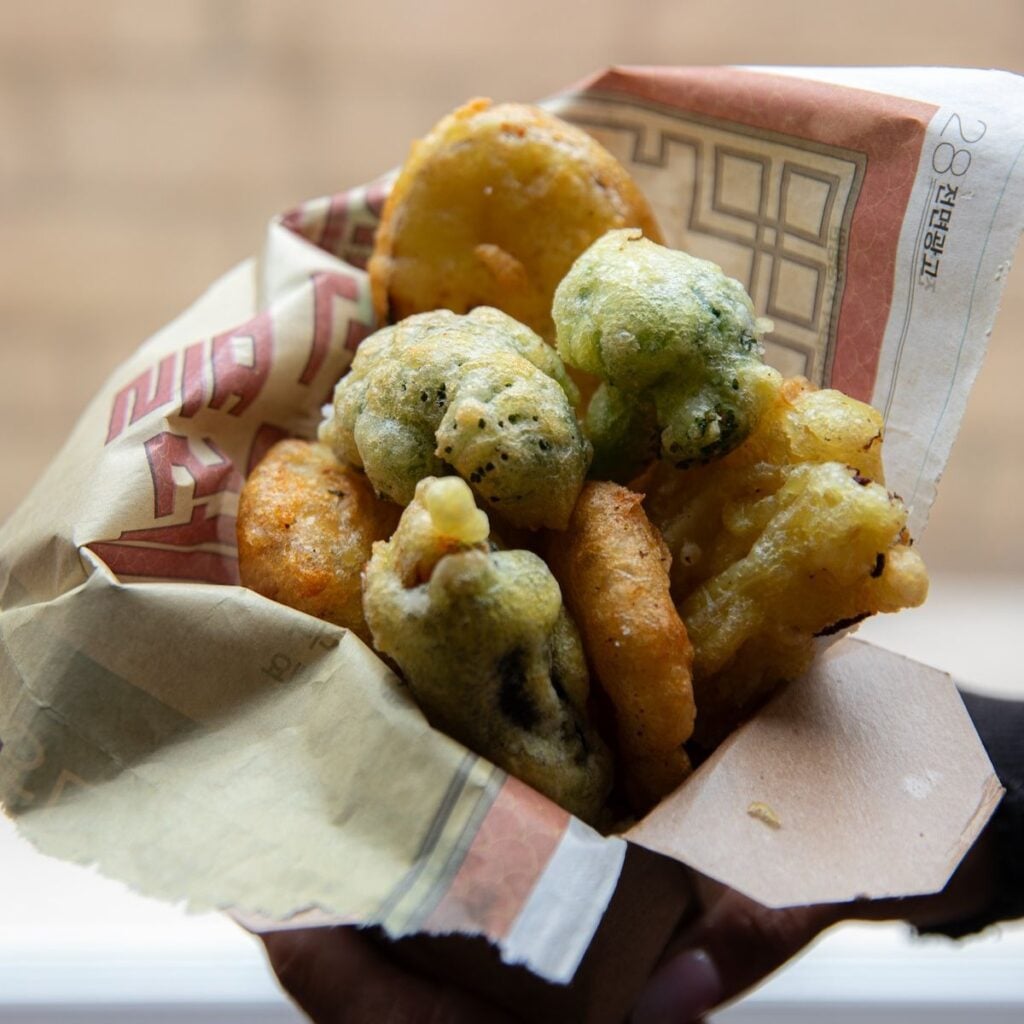 The Korean Vegan: HomeMade 47 A hand holds a paper wrap filled with assorted vegetable fritters, including golden-brown and green pieces, suggesting a mix of deep-fried snacks—perfect bites to enjoy while browsing your latest Book PreOrder against a blurred background.
