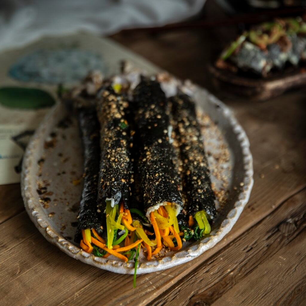 The Korean Vegan: HomeMade 51 A close-up of a rustic plate with seaweed rolls filled with julienned carrots and vegetables, sprinkled with sesame seeds, on a wooden table—perfect for your next Book PreOrder snack inspiration.