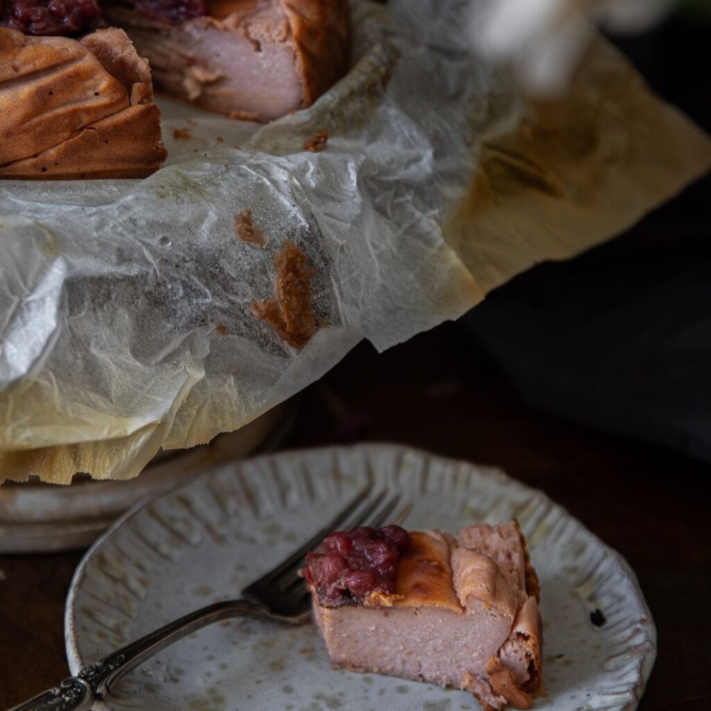 The Korean Vegan: HomeMade 45 A slice of berry-topped cheesecake on a ceramic plate with a fork, in front of the larger cheesecake resting on parchment paper. The scene is dimly lit, highlighting the dessert’s creamy texture—perfect for your next Book PreOrder snack.