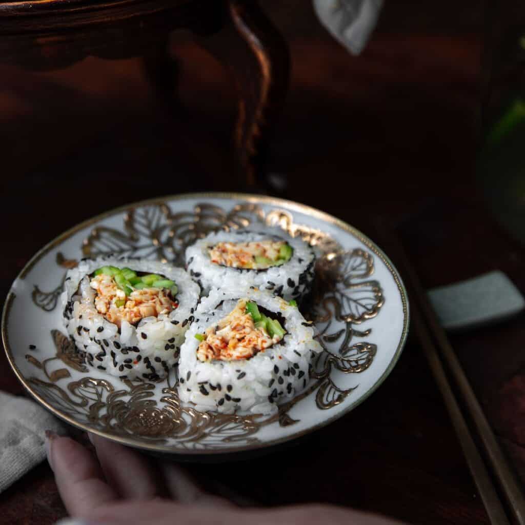The Korean Vegan: HomeMade 48 A decorative plate with three sushi rolls filled with shredded crab, avocado, and rice, sprinkled with black sesame seeds—a perfect setting to enjoy while browsing your next Book PreOrder. Chopsticks and a hand rest nearby on a dark wooden surface.