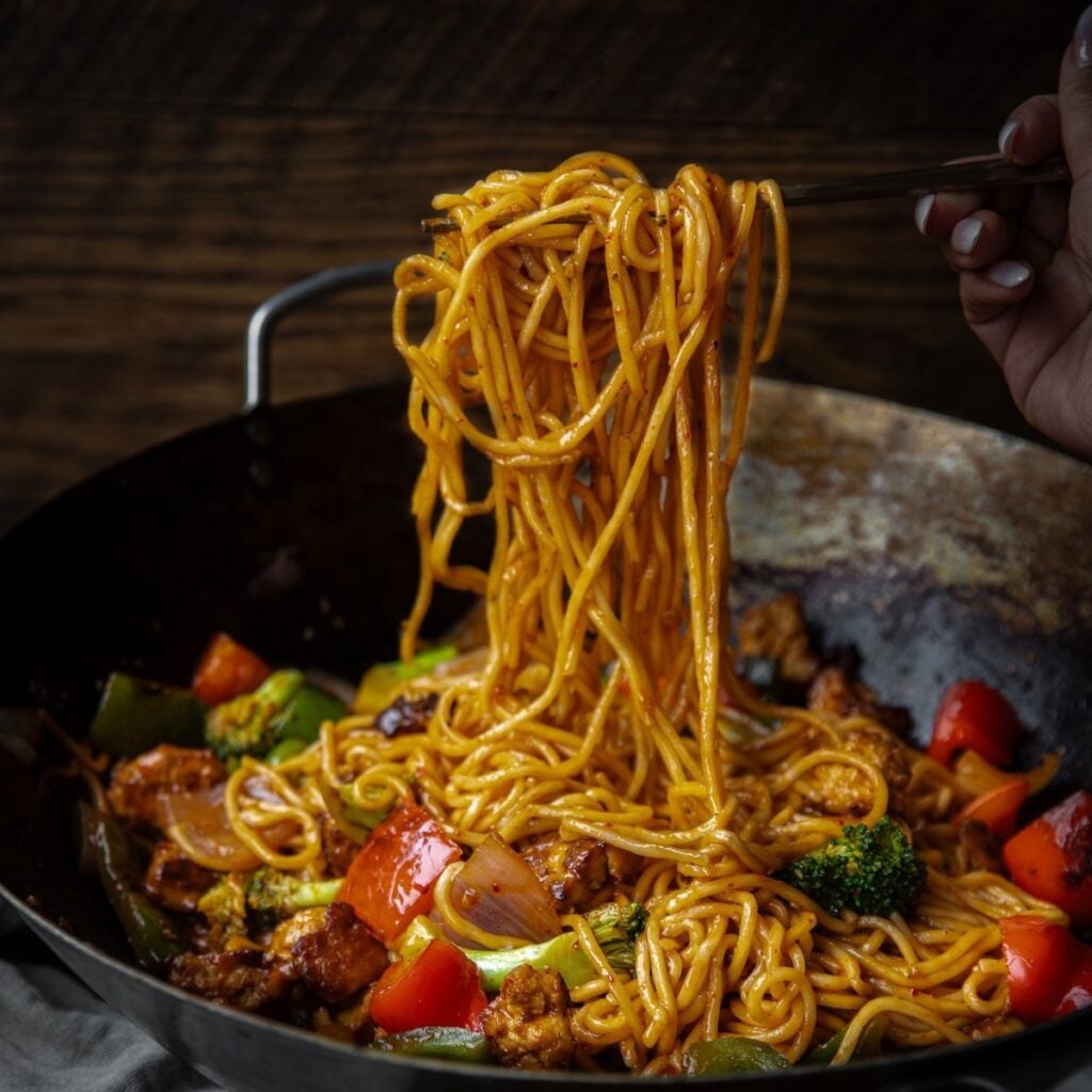 The Korean Vegan: HomeMade 46 A hand holds chopsticks lifting a portion of saucy noodles from a wok filled with noodles, broccoli, bell peppers, onions, and protein—perfect fuel while browsing the Book PreOrder Test on your favorite dark wooden table.