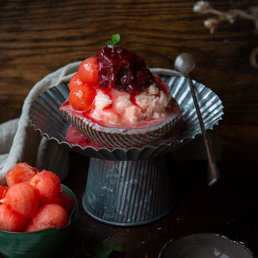 The Korean Vegan: HomeMade 49 A dessert featuring scoops of pink ice cream and red melon balls, topped with berry sauce and a mint leaf, served in a shell-shaped dish with a metal spoon—a treat worth savoring while you browse your next Book PreOrder.