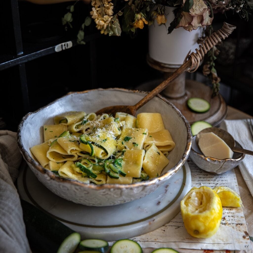 The Korean Vegan: HomeMade 52 A rustic bowl filled with wide pasta ribbons, zucchini, and grated cheese sits beside a Book PreOrder card, with a wooden spoon resting inside. Nearby are a halved lemon, cheese wedge, and zucchini slices on a textured surface.