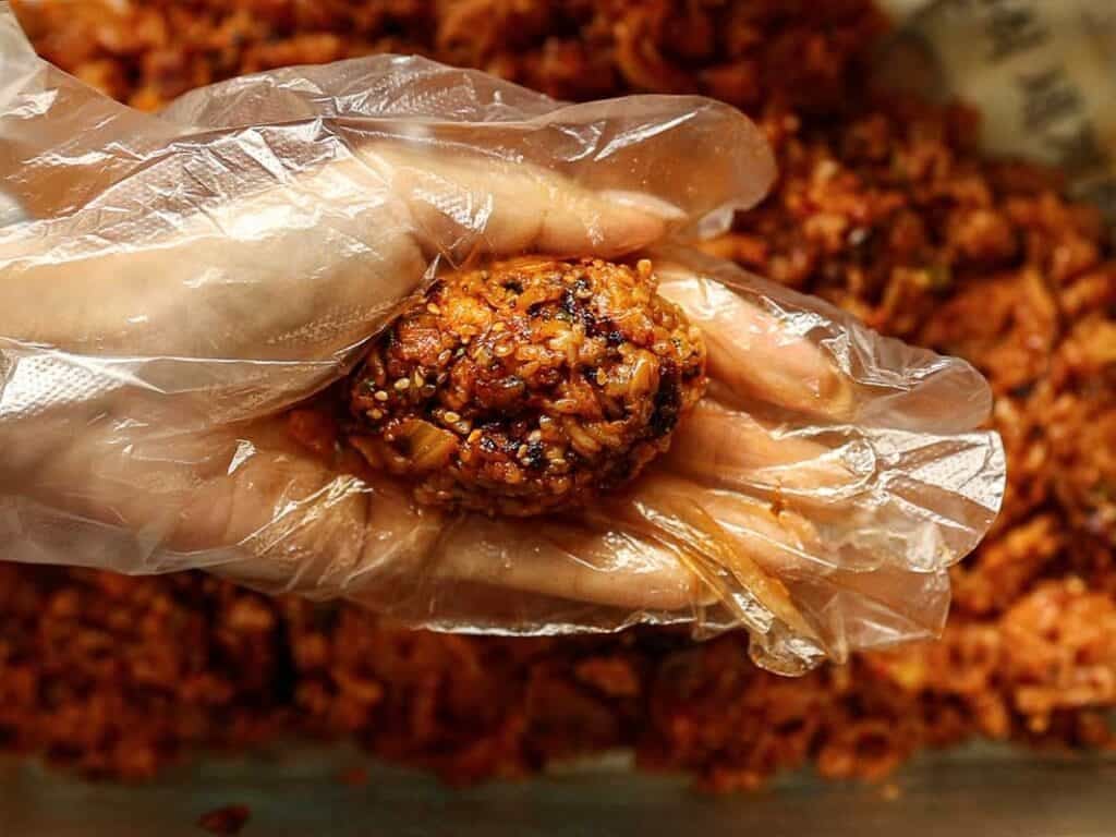 Delicious Supplì Inspired Kimchi Fried Rice Balls. 21 A hand wearing a clear plastic glove holds a ball of spicy, seasoned rice mixture, with more of the mixture visible in the background.