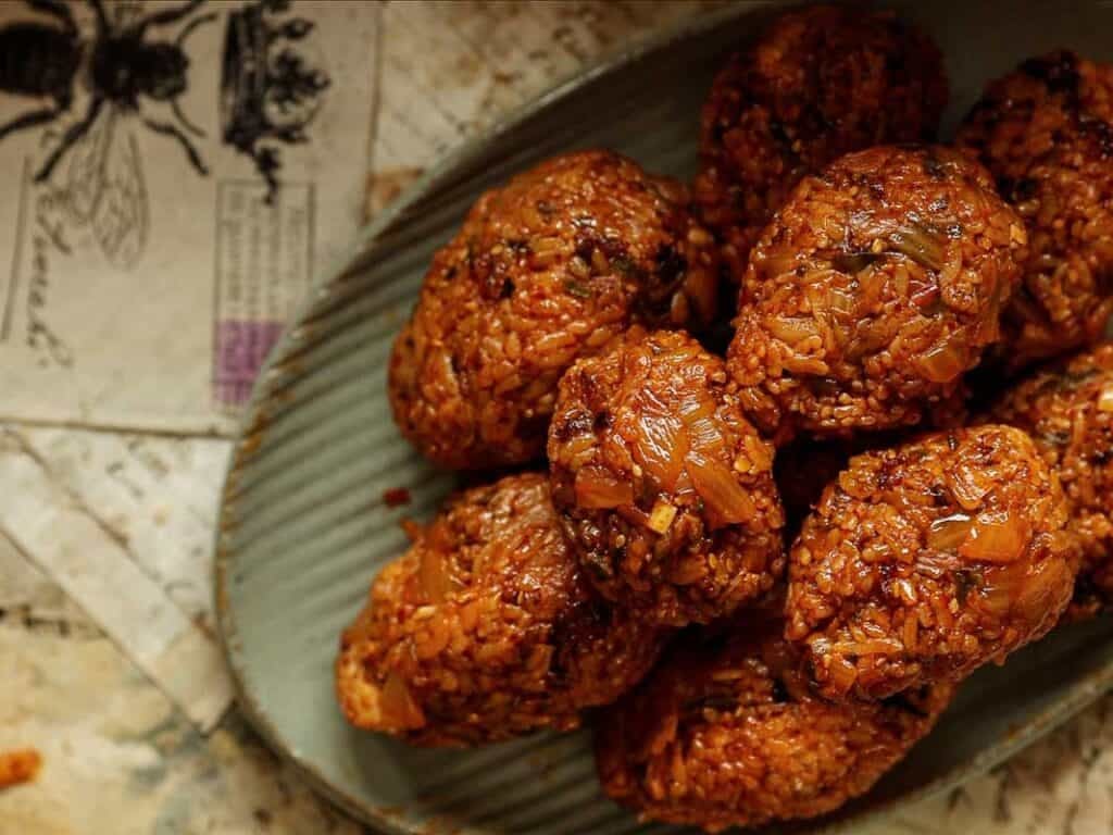 Delicious Supplì Inspired Kimchi Fried Rice Balls. 22 A close-up of oval-shaped, reddish-brown rice balls stacked on a gray plate, with some vintage papers partially visible beneath the plate. The rice balls have a glossy, textured surface.