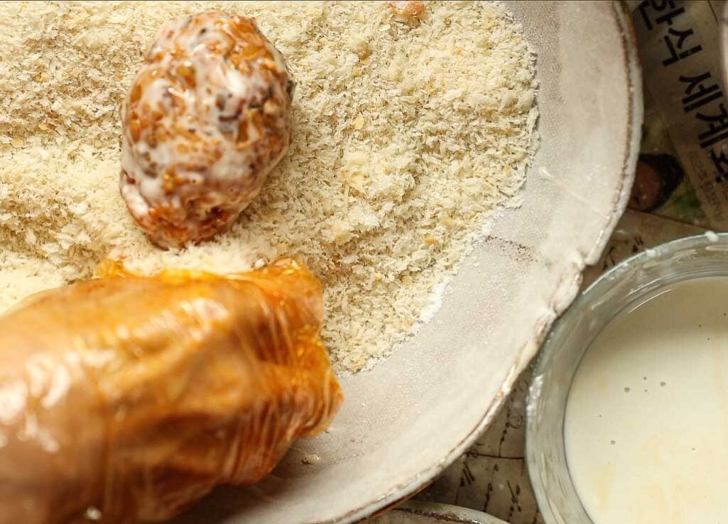Delicious Supplì Inspired Kimchi Fried Rice Balls. 23 A gloved hand coats a ball of food in breadcrumbs, with a bowl of batter and newspapers visible nearby.