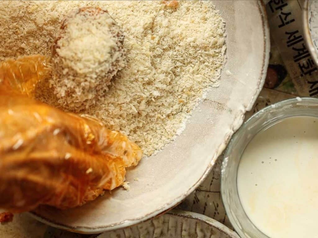 Delicious Supplì Inspired Kimchi Fried Rice Balls. 24 A hand in an orange glove coats food in breadcrumbs in a shallow bowl, with a bowl of liquid batter nearby.
