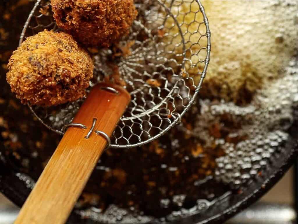 Delicious Supplì Inspired Kimchi Fried Rice Balls. 25 A close-up of two golden-brown, crispy fried balls being lifted from hot oil in a pan with a wire skimmer. Bubbles and oil can be seen in the background.