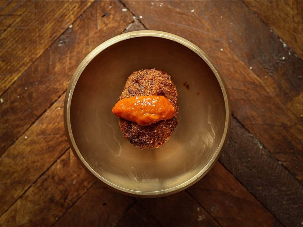 Delicious Supplì Inspired Kimchi Fried Rice Balls. 26 A single, round kimchi fried rice ball topped with a dollop of orange sauce sits in the center of a brass bowl on a rustic wooden table.