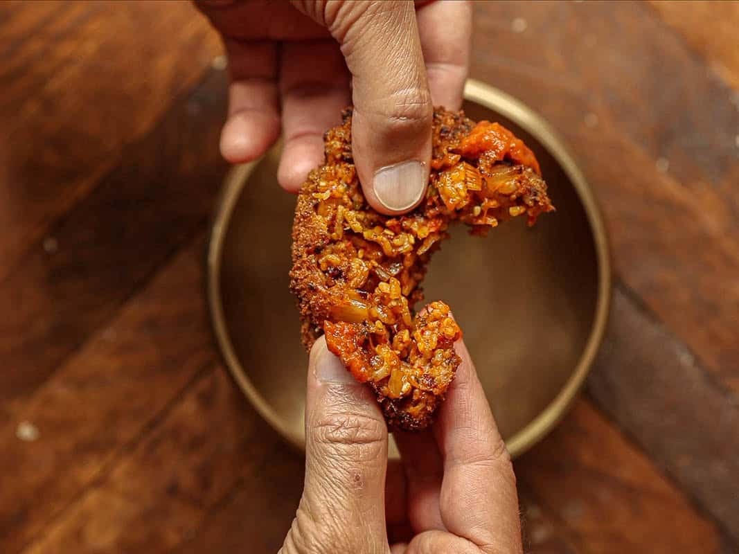 Delicious Supplì Inspired Kimchi Fried Rice Balls. 20 Close-up of two hands breaking apart a crispy, golden-brown kimchi fried rice ball with a visible vegetable filling over a round, empty bowl on a wooden surface.