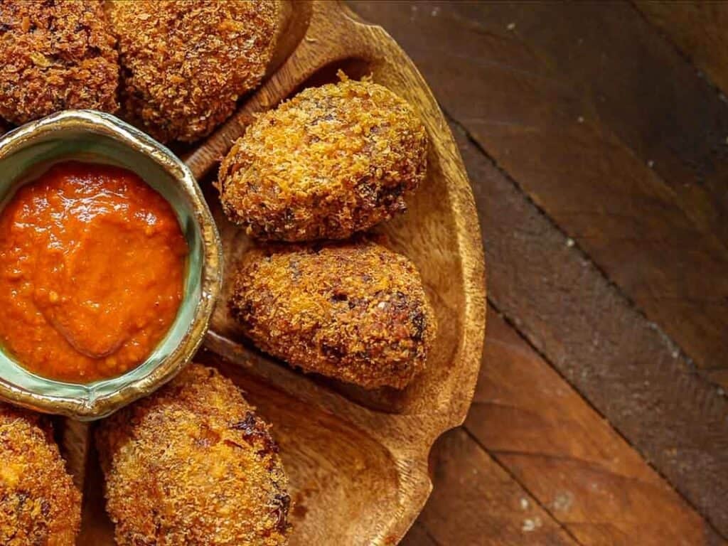 Delicious Supplì Inspired Kimchi Fried Rice Balls. 10 A wooden plate with crispy, breaded kimchi fried rice balls arranged around a small bowl of red dipping sauce, set on a rustic wooden table.