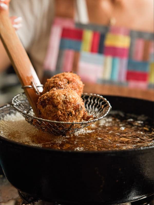 Delicious Supplì Inspired Kimchi Fried Rice Balls. 18 A person uses a slotted fryer spoon to lift two golden-brown, breaded balls from hot oil in a black pan. The background shows someone wearing a colorful, patterned apron.
