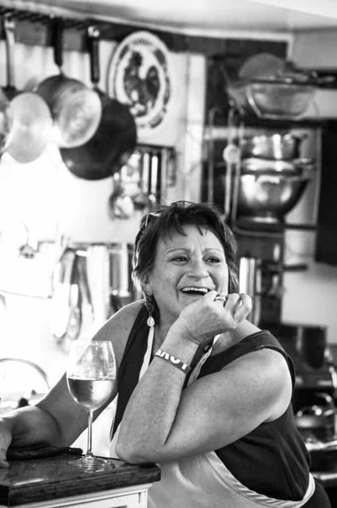 Delicious Supplì Inspired Kimchi Fried Rice Balls. 13 A woman wearing an apron sits at a kitchen counter, smiling and laughing with her chin resting on her hand. A glass of white wine is in front of her, and pots and kitchenware hang in the background. The photo is in black and white.