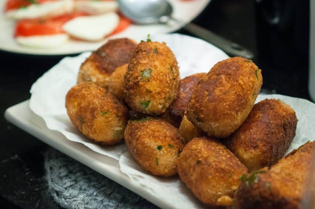 Delicious Supplì Inspired Kimchi Fried Rice Balls. 12 A plate of golden-brown croquettes sits on a white napkin, garnished with herbs. In the background, a spoon and a dish with sliced tomatoes and mozzarella are visible.