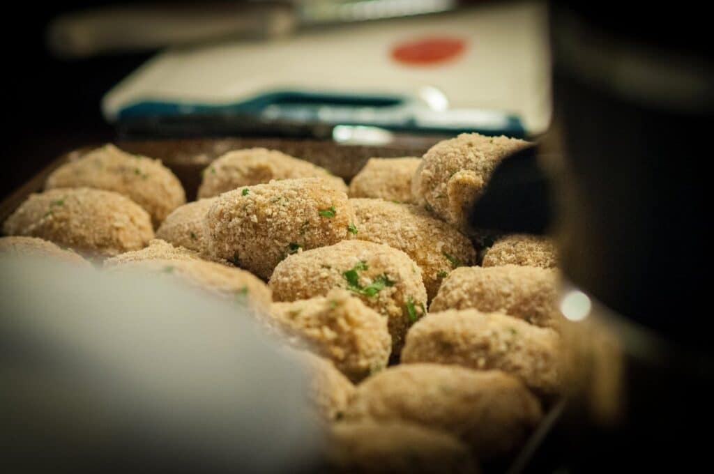 Delicious Supplì Inspired Kimchi Fried Rice Balls. 11 A close-up view of a tray filled with breaded, oval-shaped croquettes garnished with bits of green herbs, ready to be cooked. The background is softly blurred.