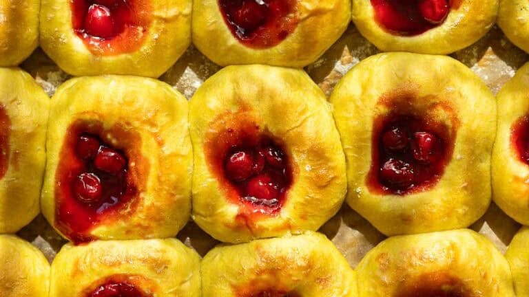 Rows of golden cherry brioche sweet roll with centers filled with glossy red cherry filling, displayed close together on a baking sheet.