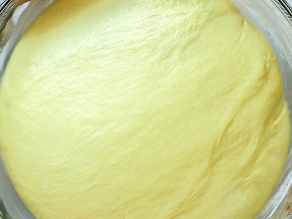 Close-up of smooth, yellow bread dough rising in a glass bowl, showing a soft and airy texture on the surface.