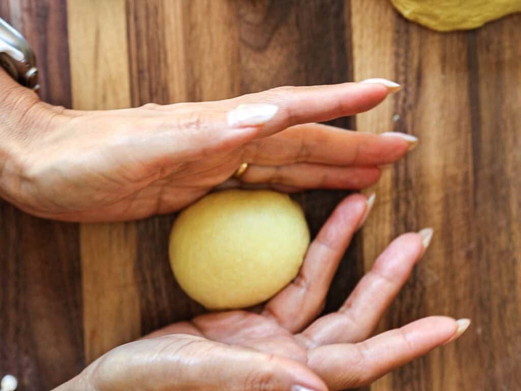 Two hands shaping a ball of dough on a wooden surface, preparing it for baking.