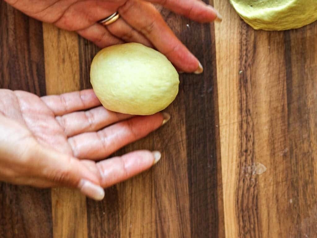 Two hands shape a piece of dough into an oval on a wooden surface, with another dough ball visible nearby.