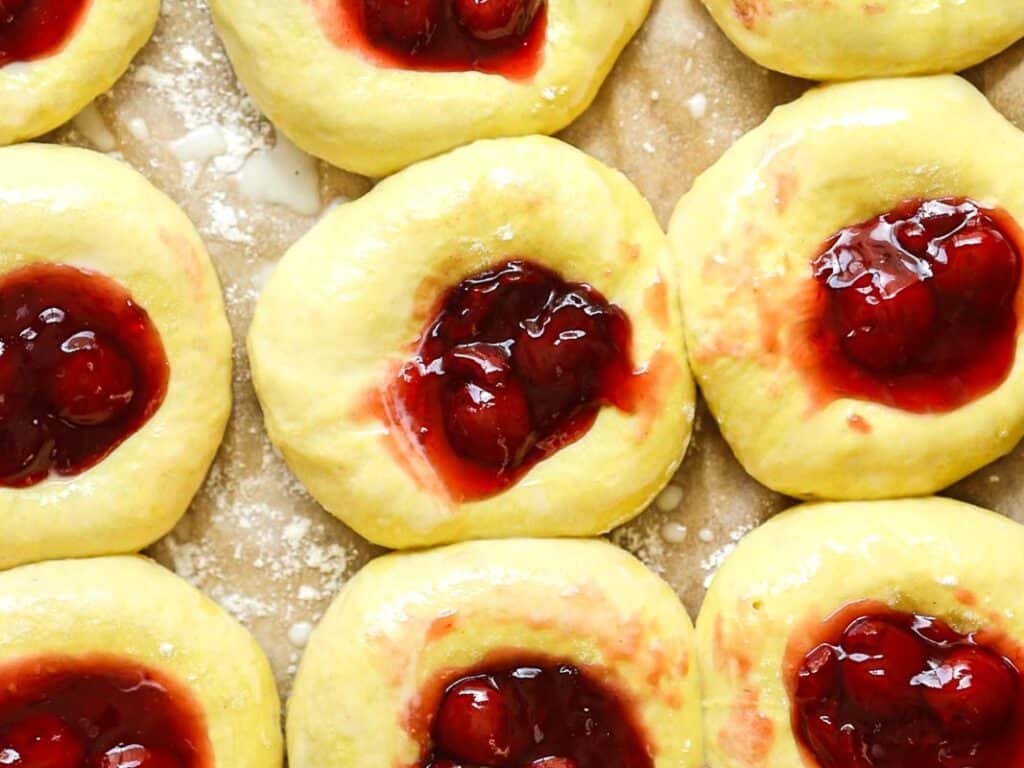Close-up of unbaked pastries with yellow dough, each topped with a dollop of cherry or berry fruit filling, arranged in rows on a floured surface.