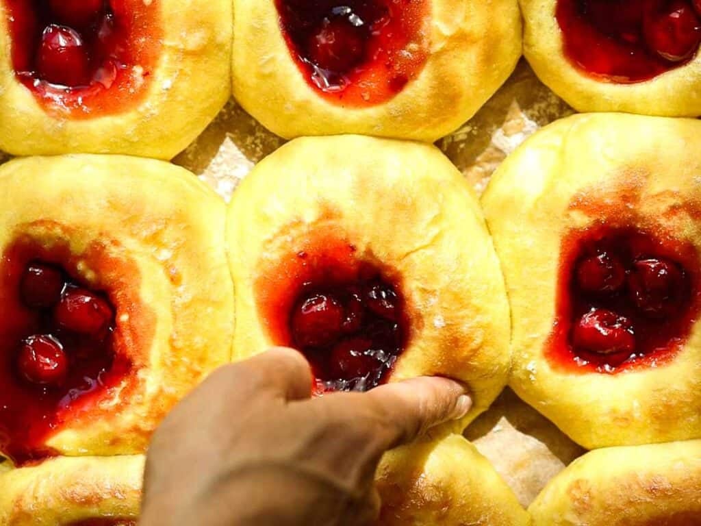 A hand presses into a soft, round pastry topped with cherry filling, surrounded by several similar pastries on a baking sheet. The pastries are golden-brown and look freshly baked.