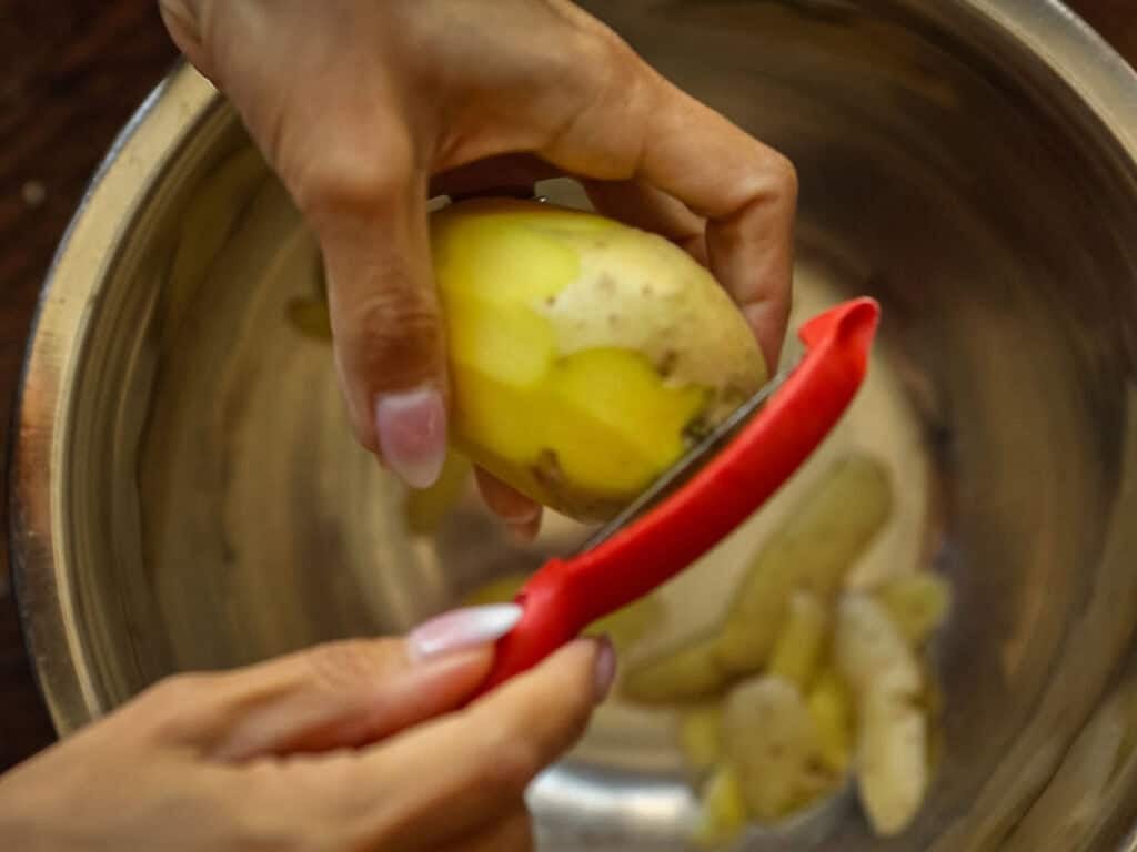 10 Tips for the Perfect Roasted Potatoes Recipe. 4 A person peels a potato with a red peeler over a metal bowl, with potato skins and pieces collected inside the bowl.