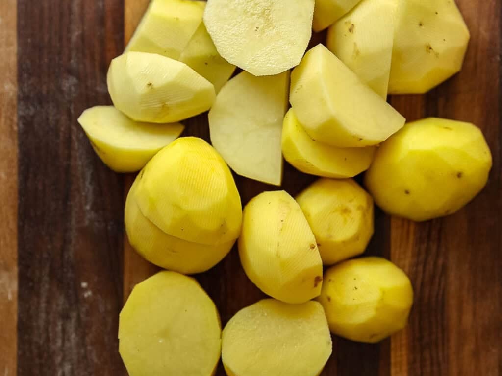 10 Tips for the Perfect Roasted Potatoes Recipe. 5 Peeled and chopped potatoes are arranged on a wooden cutting board. The potatoes are cut into halves and chunks, showing their yellow-white interior.