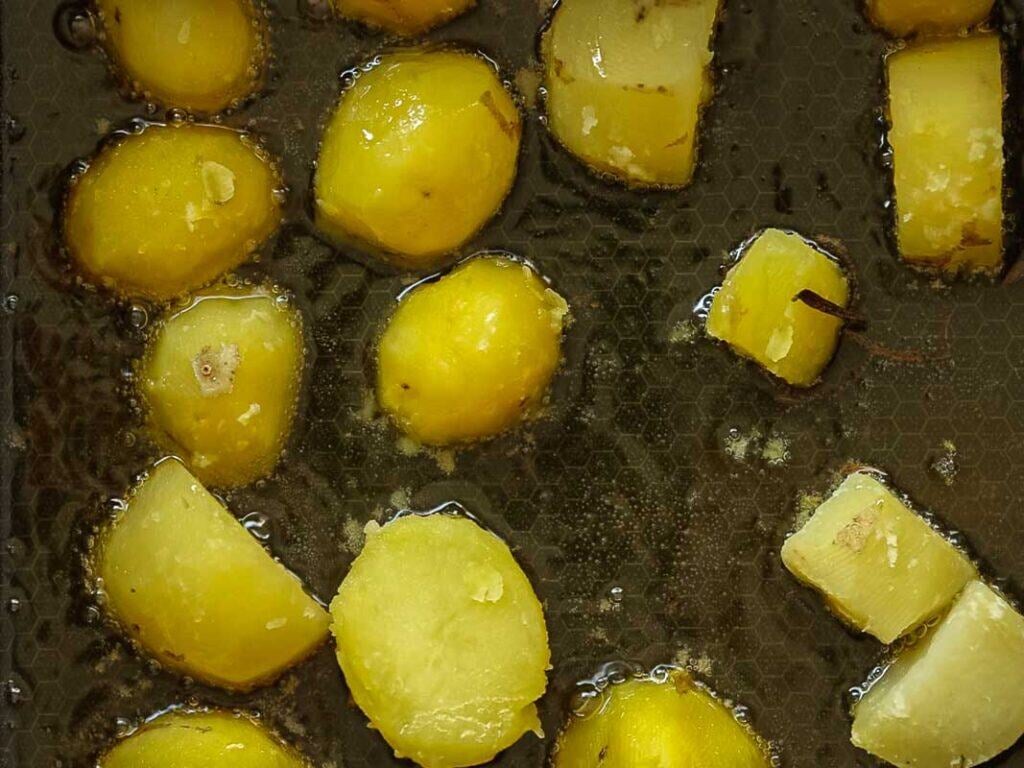 10 Tips for the Perfect Roasted Potatoes Recipe. 9 Close-up of sliced potatoes being fried in oil on a dark, textured surface. The potato pieces are golden yellow and appear partially cooked, with oil bubbling around them.