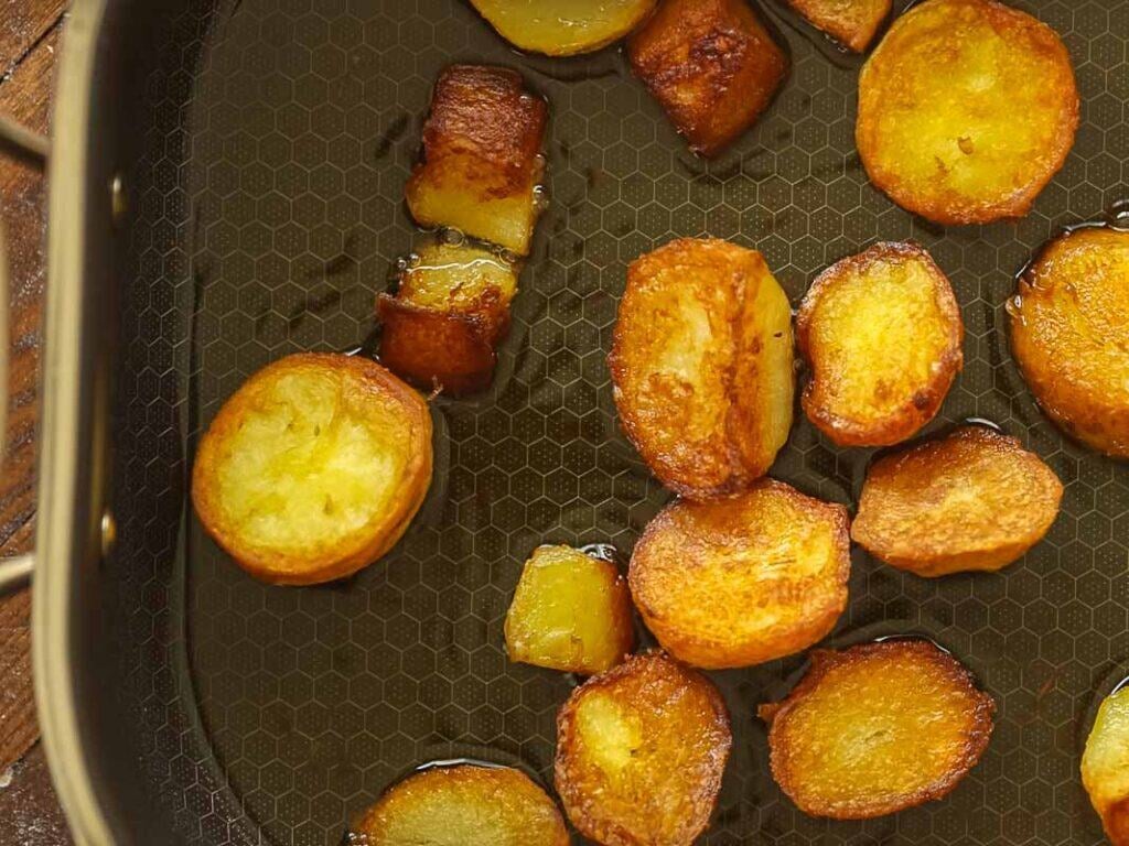 10 Tips for the Perfect Roasted Potatoes Recipe. 20 Golden brown roasted potatoes sizzling in oil on a hexagon-patterned nonstick pan. Some pieces are round and others are cubed, all with crispy edges. The pan surface is partially visible.