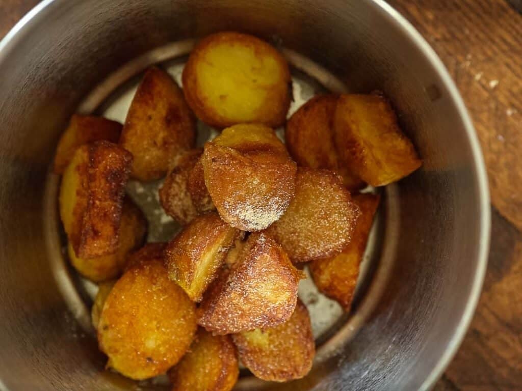 10 Tips for the Perfect Roasted Potatoes Recipe. 10 Golden, crispy roast potatoes are piled in a metal pot on a wooden surface. The potatoes have a browned, crunchy exterior and a soft, yellow interior, with a dusting of salt visible on top.