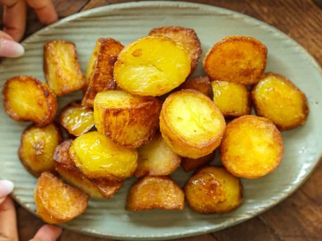 10 Tips for the Perfect Roasted Potatoes Recipe. 2 A plate of golden-brown roasted potatoes is arranged on an oval dish, with crispy edges and a shiny, caramelized surface. Two hands are holding the sides of the plate on a wooden table.
