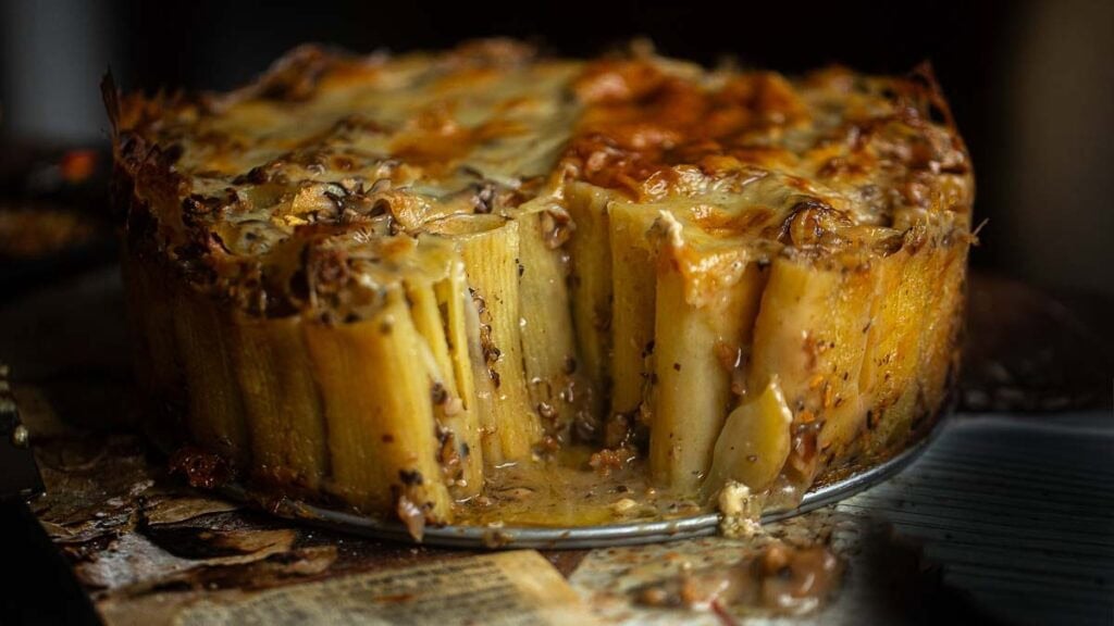 A close-up of a baked rigatoni pie with rigatoni standing upright, covered in melted cheese and sauce, with a wedge removed to reveal the inside. The pie sits on a rustic surface with some sauce spilled around.