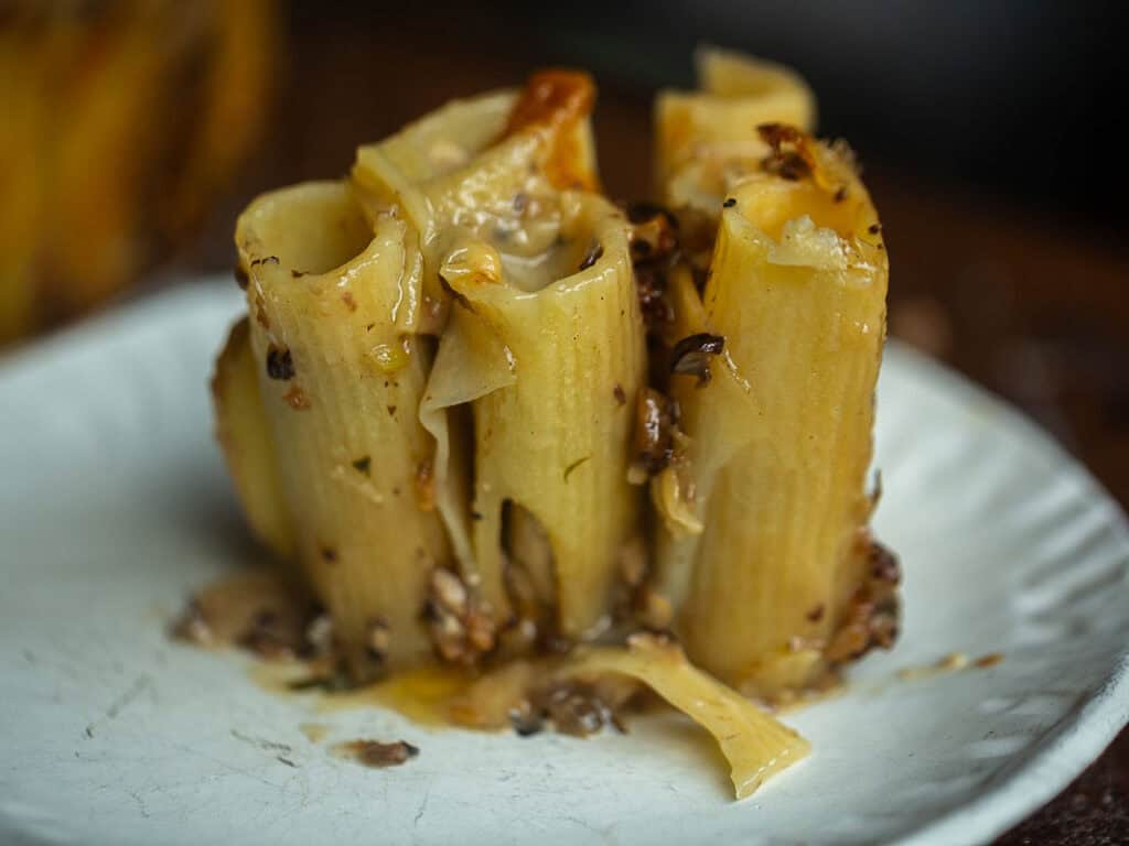 Cheesy Mushroom Rigatoni Pie - Easy Vegan Recipe. 55 A close-up of a serving of baked rigatoni pie pasta standing upright and filled with a cheesy, mushroom mixture, presented on a white plate. The pasta is golden brown with melted cheese and pieces of mushroom visible.