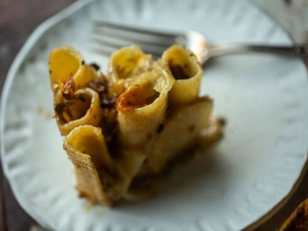 Cheesy Mushroom Rigatoni Pie - Easy Vegan Recipe. 57 A close-up of a portion of baked rigatoni pie with cheese and herbs on a white plate. A fork rests behind the pasta, and the dish sits on a rustic wooden surface.
