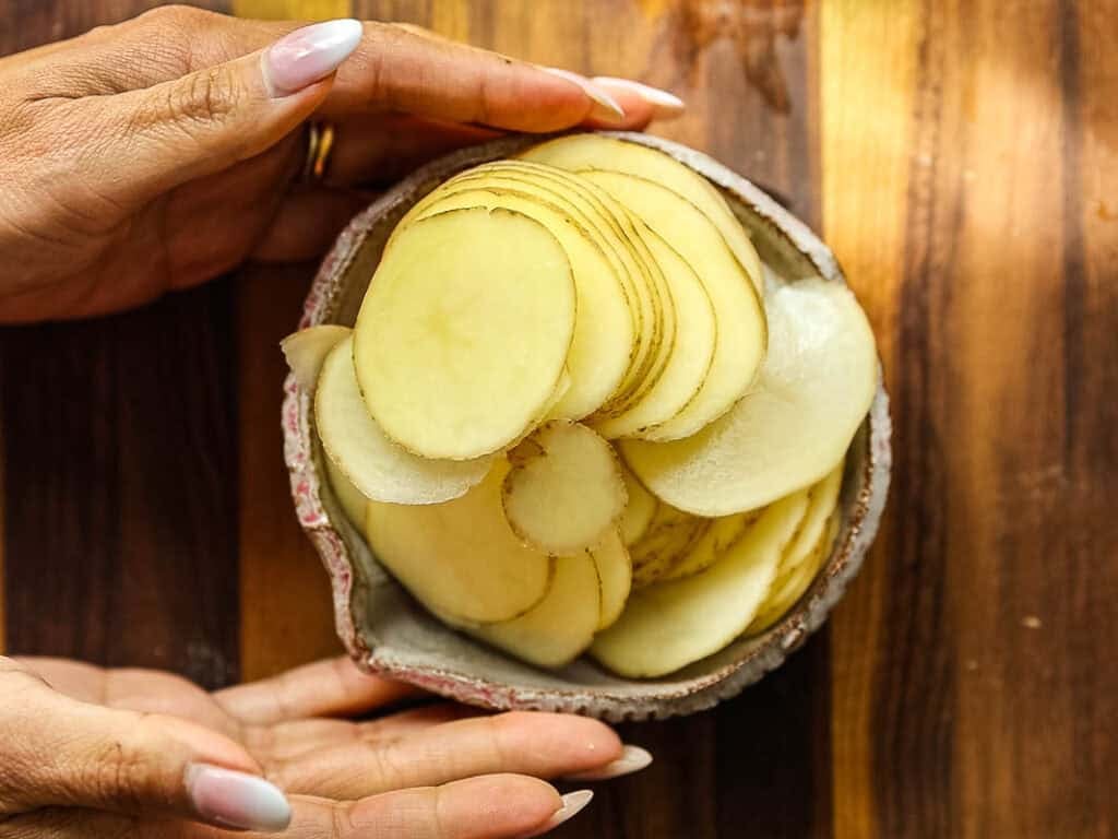 Cheesy Mushroom Rigatoni Pie - Easy Vegan Recipe. 13 A bowl filled with thinly sliced raw potatoes is held gently by two hands over a wooden surface.