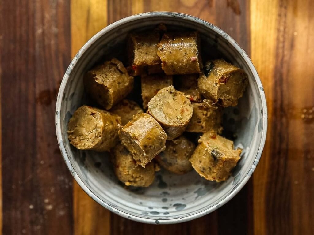 Cheesy Mushroom Rigatoni Pie - Easy Vegan Recipe. 11 A ceramic bowl filled with several pieces of sliced vegan sausage, placed on a wooden surface with striped patterns.