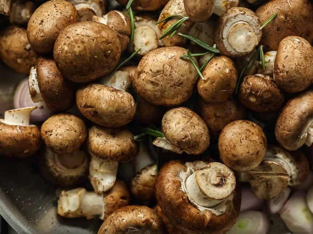 Cheesy Mushroom Rigatoni Pie - Easy Vegan Recipe. 22 A close-up of fresh brown mushrooms with sprigs of rosemary scattered among them. The mushrooms have visible stems and some sliced caps, creating a rustic, earthy appearance.
