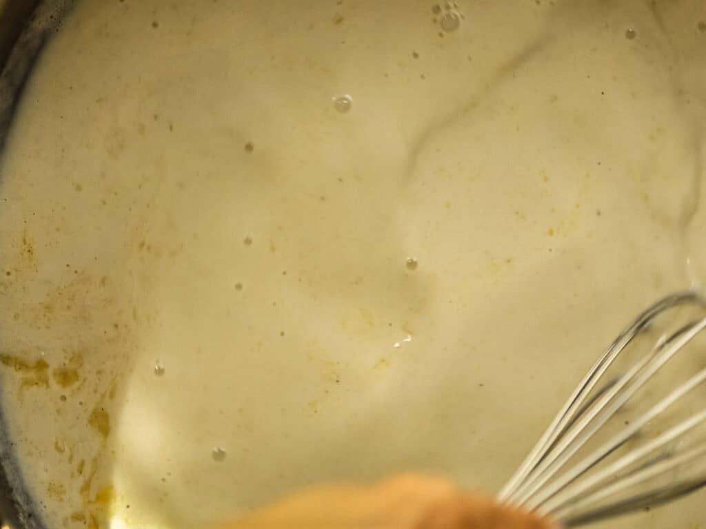 Cheesy Mushroom Rigatoni Pie - Easy Vegan Recipe. 32 A close-up of a whisk stirring a creamy, foamy liquid in a pot, with some bubbles and a light yellow tint visible on the surface.