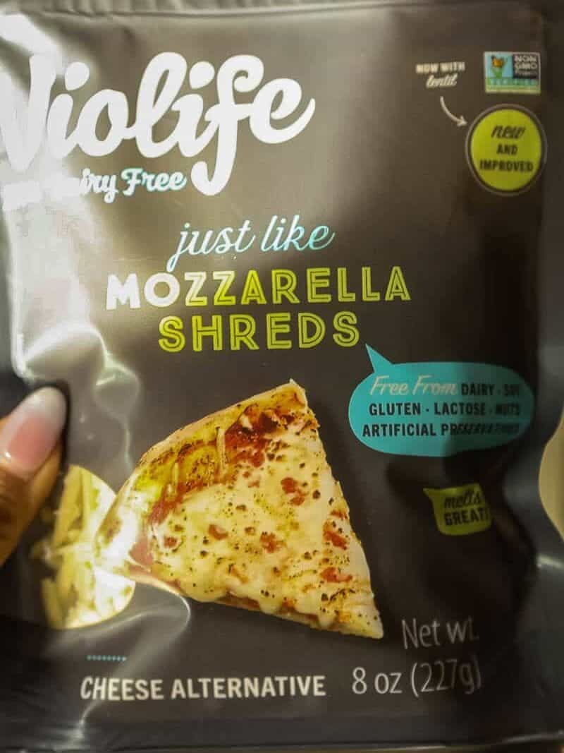 Cheesy Mushroom Rigatoni Pie - Easy Vegan Recipe. 10 A hand holds a pack of Violife Just Like Mozzarella Shreds, a dairy-free cheese alternative. The packaging shows a slice of pizza and highlights that it is free from dairy, gluten, lactose, soy, and preservatives.